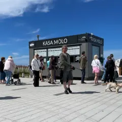 Ludzie stoją w kolejce do kasy na molo w Kołobrzegu. W tle czarny budynek z napisem "KASA MOLO".