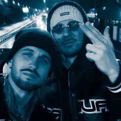 Dwóch mężczyzn w czapkach i okularach przeciwsłonecznych, jeden pokazuje gest ręką. W tle nocna droga, jakby pezet obejmuję noce.