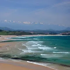 Amplias playas de arena dorada y aguas turquesas, con olas rompiendo suavemente. Al fondo, colinas verdes y montañas nevadas, un paisaje idílico de playas cerca de Comillas.