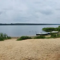 Piękny, piaszczysty brzeg jeziora w Poraju. Kilka łódek czeka na wodzie, otoczone zielenią.