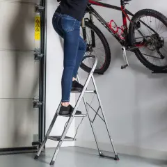 Osoba wspina się po drabince aluminiowej 11 szczebli, by sięgnąć po rower wiszący na ścianie.
