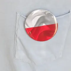 Okrągła przypinka z polską flagą wpięta w kieszeń jasnoniebieskiej koszuli.