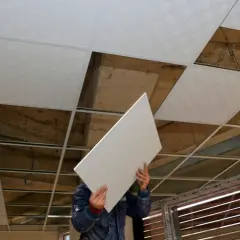 Pracownik montuje płyty do ocieplenia sufitu podwieszanego. Widać odsłonięte elementy konstrukcji i stare betonowe strop.