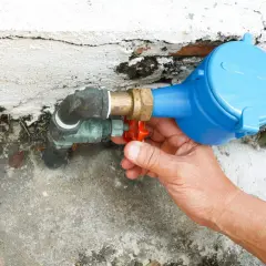 Main d'une personne actionnant le robinet rouge du tuyau d'arrivée d'eau de la maison, juste avant le compteur bleu.