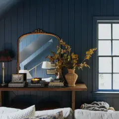 A rustic style console table with a lamp, books, and a vase of autumn leaves sits against a dark blue paneled wall, reflecting in an ornate mirror.