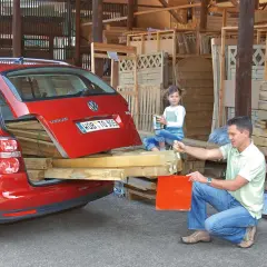 Familie belädt roten Kombi mit Holzbrettern. Die Heckklappe ist offen, die **Ladung nach hinten** überstehend. Ein Mann und eine Frau packen an, ein Kind schaut zu.