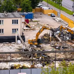 Dwa koparki pracują przy rozbiórce budynku. W tle widać ruchliwą ulicę i zaparkowane samochody.