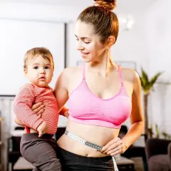 Młoda mama z dzieckiem na ręku mierzy talię centymetrem. Pokazuje, jak schudnąć po ciąży, łącząc macierzyństwo z dbaniem o formę.