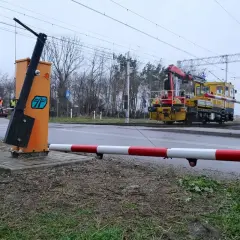 Żółta lokomotywa z dźwigiem zbliża się do przejazdu kolejowego bez zapór. Na pierwszym planie pomarańczowy słup z podniesionym ramieniem.