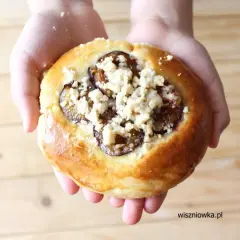 Drożdżówka z śliwkami i kruszonką trzymana w dłoniach na drewnianym tle.