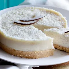 Delizioso cheesecake al cocco, ricoperto di cocco grattugiato e decorato con scaglie di cocco fresco. Una fetta è già stata tagliata, invitando a gustare questo dolce tropicale.