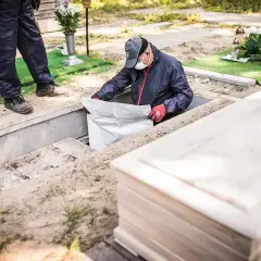 Ekshumacja zwłok po 40 latach. Pracownik w masce i rękawiczkach wyciąga worek z grobu. Obok leży nowa trumna.