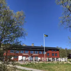 STF Vålådalen fjällstation i solsken med svensk flagga. Träd med gröna blad ramar in den röda byggnaden.