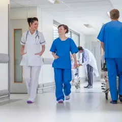 In un corridoio ospedaliero, personale medico si muove con urgenza. Un uomo spinge una barella, mentre due infermiere camminano velocemente, forse a seguito di un evento avverso.