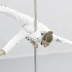 Dziewczyna w białym stroju z gwiazdami wykonuje akrobatyczną figurę na rurze. Czy pole dance to grzech? Ta młoda sportsmenka udowadnia, że to sztuka.