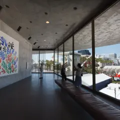 Inside a modern art museum, a colorful Matisse cutout hangs on a concrete wall. Visitors admire the art, enjoying the view of the city skyline.