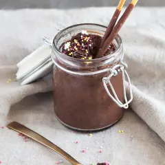 Un pot de mousse au chocolat mascarpone, garni de vermicelles colorés et de bâtonnets au chocolat. Une cuillère dorée repose à côté.