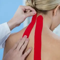 Fizjoterapeuta aplikuje czerwony taping medyczny na kark pacjentki, wspierając mięśnie i poprawiając krążenie.