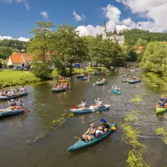 Wielu ludzi płynie kajakami i pontonami po rzece w Czechach, podziwiając malownicze widoki i zabytkowy kościół na wzgórzu.