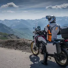 Motocyklista w kasku i kurtce podziwia górską panoramę Alp z krętą drogą w dole.