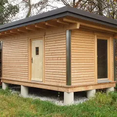 Moderne gartenhaus ideen: ein Holzhaus auf Betonpfeilern mit Pultdach, Holztür und großem Fenster. Daneben ein Holzstapel.