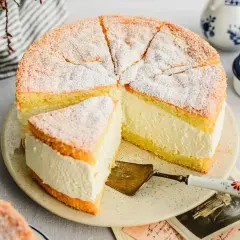 Saftige Quark Sahne Torte, mit Puderzucker bestäubt, auf einem Teller mit Tortenheber. Daneben Zitronen und eine Kanne.