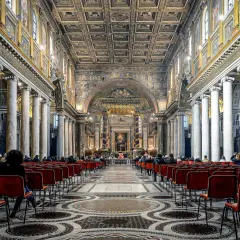 Intérieur de Sainte-Marie-Majeure, Rome. Des fidèles assis sur des chaises rouges, sous des colonnes imposantes et un plafond orné.
