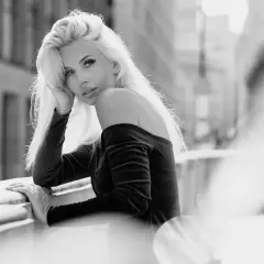 Elegancki portret w plenerze. Kobieta z blond włosami opiera się o balustradę, patrząc w obiektyw.
