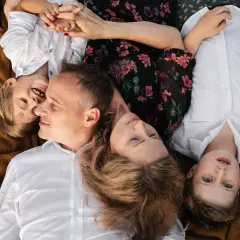 Rodzina leży na kocu, tworząc śmieszną sesję rodzinną. Dzieci śmieją się, a rodzice patrzą na siebie z miłością.