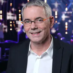 Uśmiechnięty Robert Janowski w okularach na tle sceny. Wiele osób zastanawia się, ile lat ma Robert Janowski, ale jego wiek nie jest widoczny na zdjęciu.