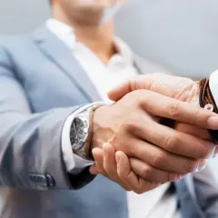Uścisk dłoni dwóch mężczyzn w garniturach, jeden z zegarkiem, drugi z bransoletką.