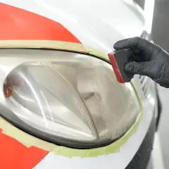 Mano con guanto nero lucida un faro auto opaco, pronto per verniciare. Nastro protettivo arancione e verde.