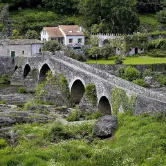 Un antiguo ponte carreira de piedra cruza un río, rodeado de casas rústicas y vegetación exuberante.