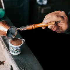 Mężczyzna nalewa parzącą się kawę z miedzianego tygielka do ozdobnej filiżanki.