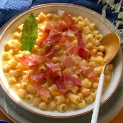 Un piatto fumante di pasta e ceci, arricchito da fette di prosciutto crudo e una foglia di alloro.