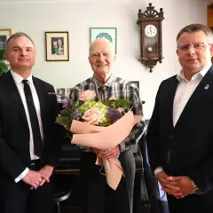 Trzech mężczyzn, jeden z bukietem kwiatów, w eleganckich strojach.