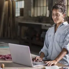 Mujer en taller revisa sus cuentas anuales en su portátil, rodeada de herramientas y madera.