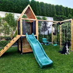 Drewniany plac zabaw z zieloną zjeżdżalnią, huśtawkami i ścianką wspinaczkową, na trawniku, z psem rasy border collie.