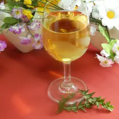 Szklanka złocistego wina z mniszka lekarskiego, otoczona sztucznymi kwiatami i liśćmi mniszka.
