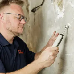 Mann mit Brille misst mit einem Gerät die Feuchtigkeit einer Kellerwand.