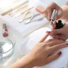 Dłoń podczas malowania paznokci ciemnym lakierem, w tle przybory do manicure.