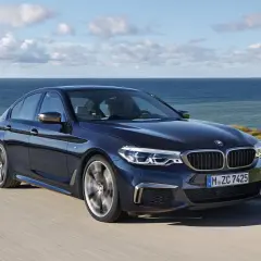 Ciemnoniebieskie BMW M550i sunie po krętej drodze nad brzegiem morza.