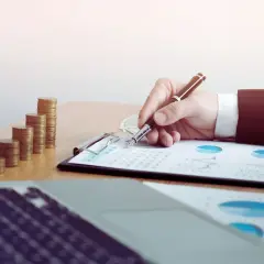 Ręka pisze na dokumentach z wykresami, obok stosy monet. Sąd może żądać wyciągu z konta w sprawach finansowych.