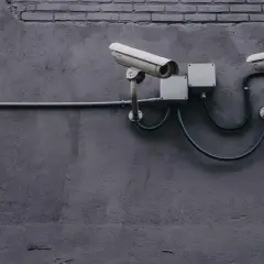 Dwie kamery monitoringu na szarej ścianie, podłączone do puszek elektrycznych. Zapewniają bezpieczeństwo i monitoring w domu.