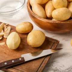 Vorbereitung für Kartoffelgerichte: Ein Messer liegt auf einem Holzbrett neben geschälten und ungeschälten Kartoffeln. Eine Schüssel mit Kartoffeln steht daneben. Wie viel Kartoffeln pro Person?