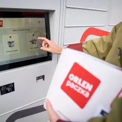 Dłoń dotyka ekranu automatu paczkowego, wybierając opcję "Nadaj przesyłkę". W ręku trzymana jest biała paczka z logo ORLEN paczka.