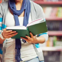 Osoba w okularach i szaliku czyta otwartą zieloną książkę na tle regałów z książkami.