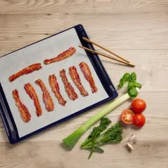 Bacon i ugn på bakplåtspapper, bredvid ligger grönsaker och vitlök.