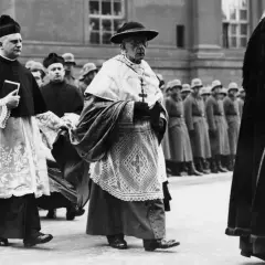 Geistliche in feierlicher Kleidung marschieren, im Hintergrund Soldaten. Die Kirchensteuer im Nationalsozialismus war ein komplexes Thema.