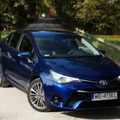 Błękitna Toyota Avensis T27 na żwirowej drodze, otoczona zielenią i drzewami.